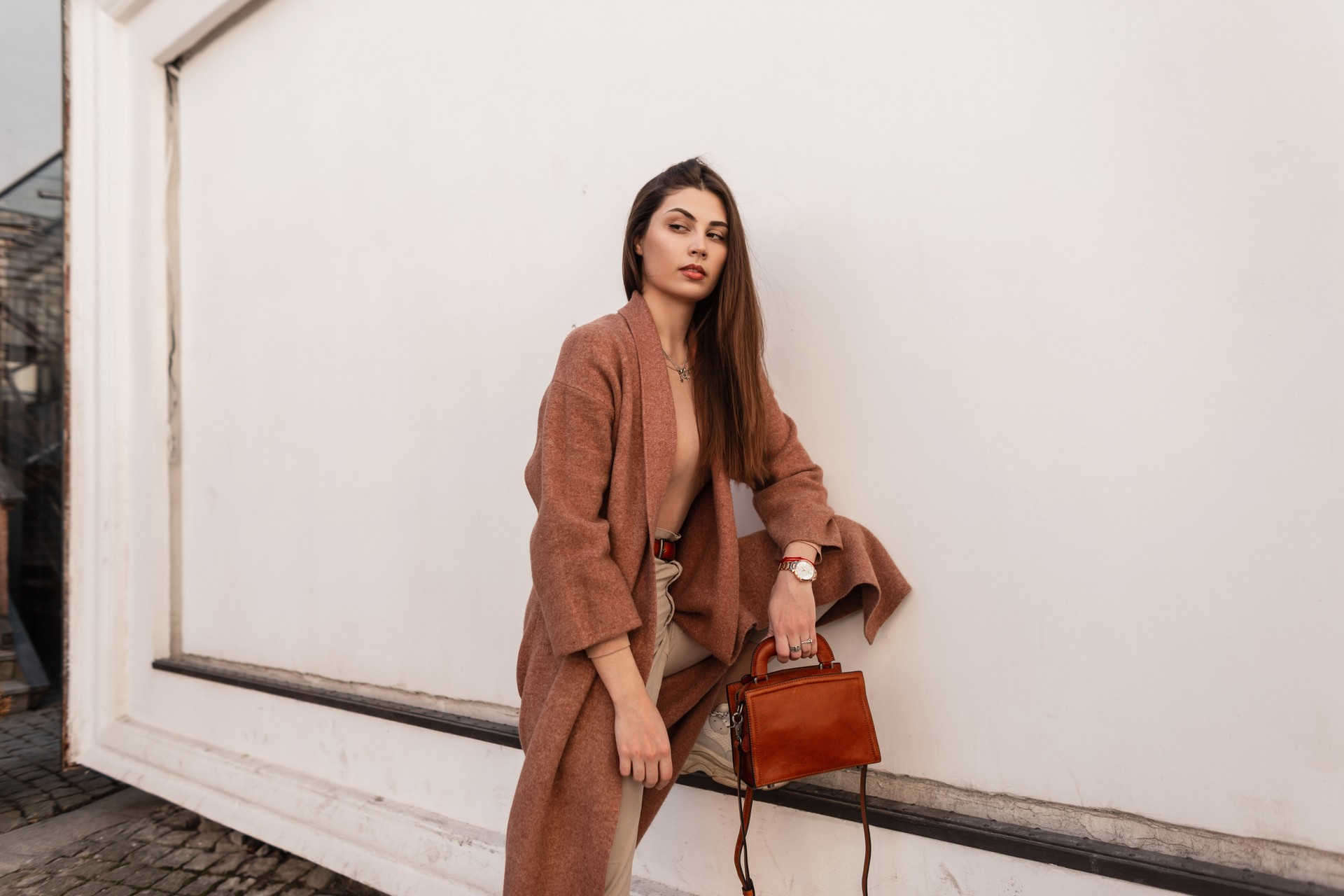 Stylish beautiful girl in fashionable outerwear stands near the wall